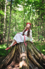 Fototapeta premium Beautiful young smiling woman wearing Slavic white dress sitting on the stump in the forest