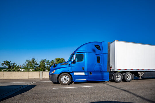 Bright blue big rig bonnet semi-truck with high cab transporting cargo in dry van semi trailer driving on the multiline highway road