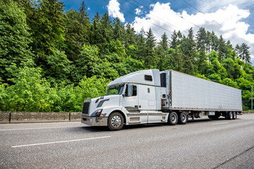 Popular comfortable big rig semi truck tractor transporting cargo in refrigerated semi trailer driving on the wide highway road with green forest on the side