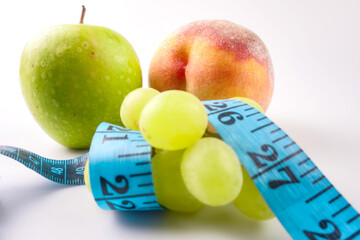 A peach, an apple and a bunch of grapes wrapped with measuring tape lie on a white background. Close up.