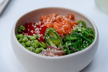 Close-up view of seafood poke bowl with salmon, rice, cucumber, radish, nori seaweed and edamame beans on white table in restaurant or sidewalk cafe. Soft focus. Copy space. Food theme.