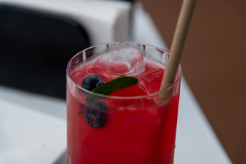 Close-up view of red lemonade in drinking glass standing on white table in sidewalk cafe or street restaurant. Ice cubes, blueberries and leaf of mint floats on top of cold drink. Summer drinks theme.
