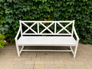 A white bench rests on a brick sidewalk near a green hedge, creating a serene spot to unwind outdoors under the sunny sky. Its a tranquil setting to relax and soak in natures beauty