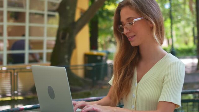 Happy female freelancer typing on laptop sitting at street cafe. Confident young woman in trendy glasses using netbook for new blog on outdoor cafe terrace. Blogger writing outdoors for blog.
