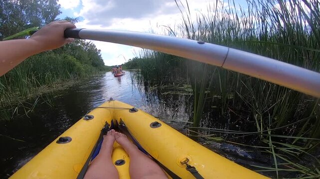 Person floating in inflatable boat along river through thickets of green reeds on sunny summer day. People kayaking along river. Man in boat rowing oars. Sport, action, activity, lifestyle. POV