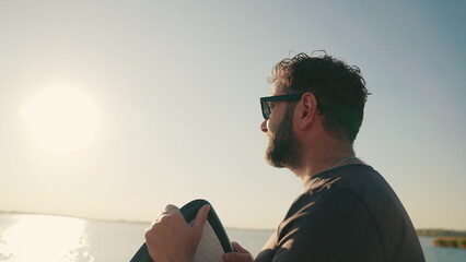Close-up portrait of a man standing on a lake pier at sunset, leaning on a waterboard. Male surfer...