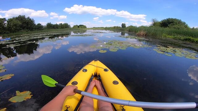 Kayaking along river through growing white lily nymph on sunny summer day. Person floating in boat on river with surface of water reflecting white clouds on calm day. Tourism. Active lifestyle. POV