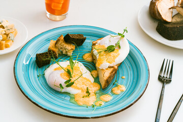 Poached eggs on toast with hollandaise sauce and crouton on a turquoise plate and slices of toasted bread on a white plate on a light background. Close-up. Selected focus.