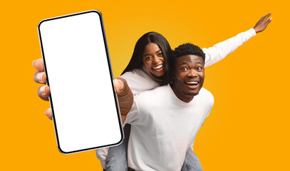 A young African American couple standing in front of a yellow background. The man is holding a smartphone in front of him, with the screen blank.