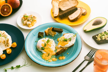 Eggs Benedict with hollandaise sauce and peas on a turquoise plate and avocado with rye toast, cheese, blood oranges and orange juice on a white table.