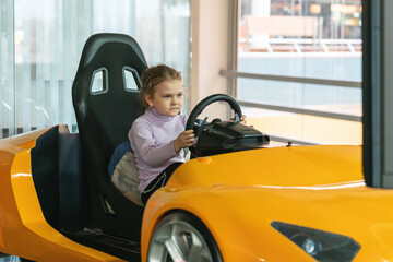 A little girl in an entertainment center with slot machines sits in a car game simulator and holds...
