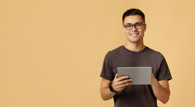 Modern work, social media and online communication. Attractive happy young male student in glasses, with smart watch typing in digital tablet, isolated on beige background, studio shot, free space
