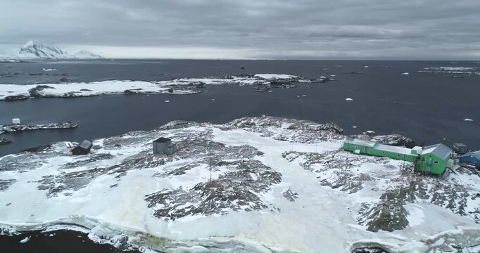 Polar station buildings on rocky island in Antarctica. Frozen coast, cold ocean, mountains in background. Winter wild nature landscape. Antarctic expeditions, travel. South Pole explore. Drone flight