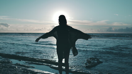 Woman raise hands up on sunset sea beach. Female silhouette stand on ocean coast, wind blow hair...