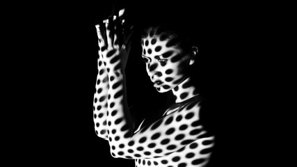 A black and white image of a woman standing in a dark studio. Her body is illuminated by a projected pattern of dots, casting an abstract design on her skin. © Prostock-studio