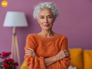A senior biracial woman with short, white hair looks thoughtful while standing in a cozy, home setting. She is wearing an orange sweater, and the background features a pink wall and a lamp.