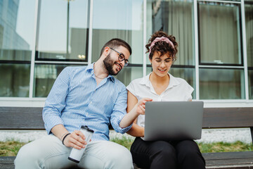 Two smiling business people working on laptop in front modern business building