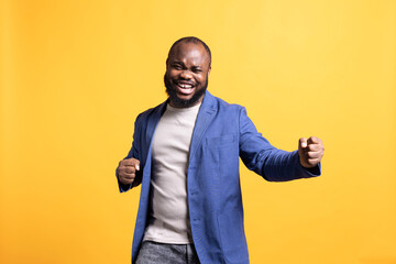 Portrait of energetic happy african american man celebrating, throwing fists in air from excitement. Overjoyed BIPOC person enjoying success, cheering and gesticulating, studio background