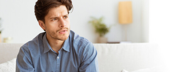 A young man sits on a white couch, looking up and to the right, lost in thought.