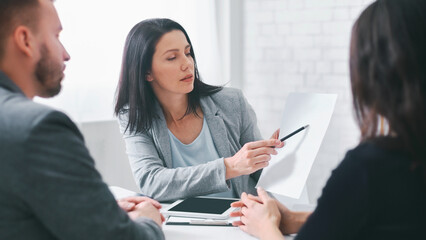 A businesswoman is presenting information to two clients in an office setting. She is pointing at a...