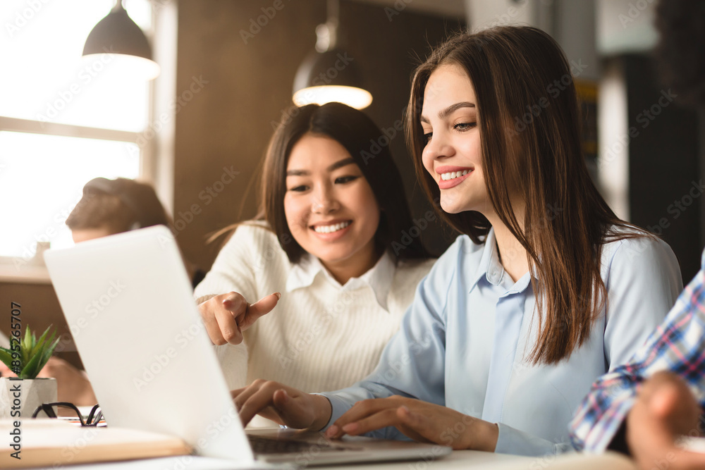 Poster two multiethnic women are seated at a desk in a well-lit library, working together on a project. one