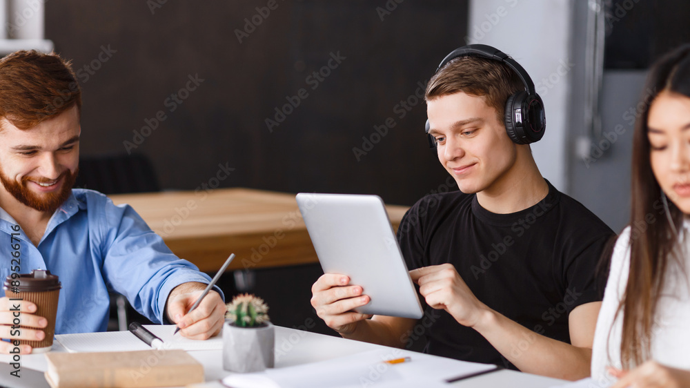 Poster A group of young adults is gathered around a table in a well-lit office space, collaborating on a project. One is smiling while holding a digital tablet, another is taking notes