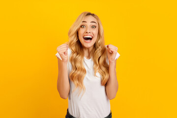 A young woman with blonde hair and a white t-shirt stands in front of a yellow background. She is...
