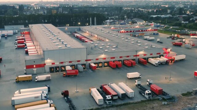 Aerial View of Bustling Logistics Park at Dusk | Illuminated Warehouses and Truck Fleet in Urban Setting