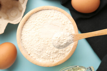 Bowl with wheat flour, spoon and eggs on blue background