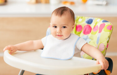 A cute baby wearing a white bib sits comfortably in a high chair in a well-lit kitchen. The baby looks curious and content while holding onto the edge of the high chair.
