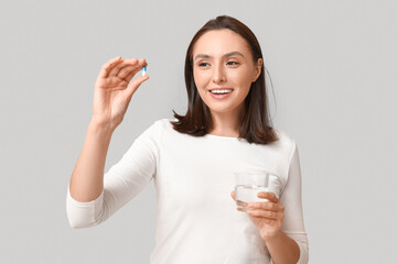 Pretty young woman with pill and glass of water on grey background