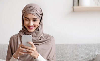 Portrait of millennial muslim girl in hijab using smartphone at home, messaging or browsing social networks, sitting on sofa in living room