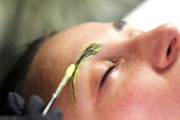 Close-up of a beautician applying a yellowish tint to a woman's eyebrows with a brush. The woman is lying down with her eyes closed, emphasizing the careful and precise application.