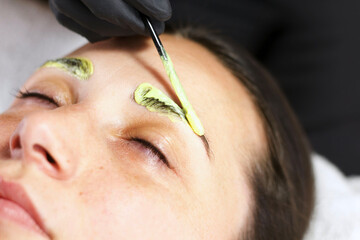 A beautician applies a yellowish tint to a woman's eyebrows using a brush. The woman is lying down with her eyes closed, receiving professional beauty treatment. - Powered by Adobe