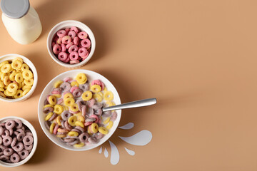 Bowls with colorful sweet cereal rings and bottle of milk on brown background