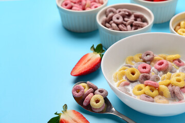 Bowls of colorful sweet cereal rings with milk and cut strawberry on blue background