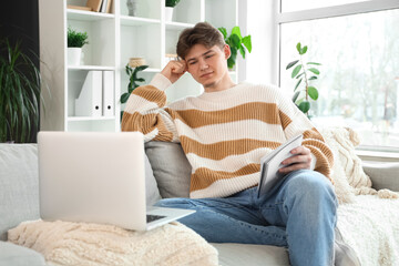 Fototapeta premium Male student with notebook and laptop studying online on sofa at home