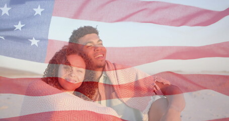 Image of american flag over happy diverse couple sitting on sunny beach embracing