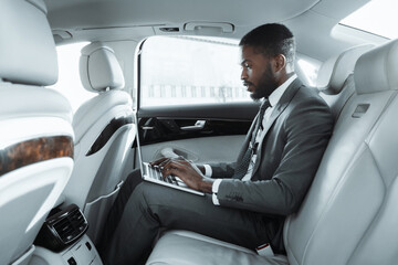 Black businessman in a suit sits in the back seat of a luxury car, working on a laptop computer. He is focused on his work, typing on the keyboard with his right hand