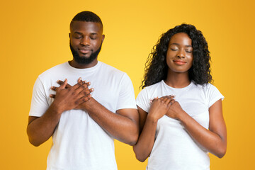 A Black couple stands with their eyes closed, holding their hands over their hearts.