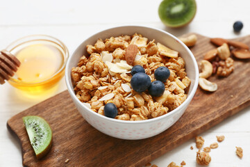 Wooden cutting board, bowl with tasty granola, nuts and berries on white wooden background, closeup