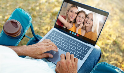 A senior man sits on a blue lawn chair outdoors and uses a laptop to have a video call with his family. He is smiling and appears happy to be connected to his loved ones