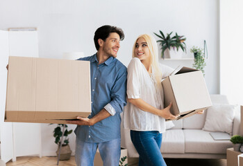 A couple smiling and holding cardboard boxes in a new house.