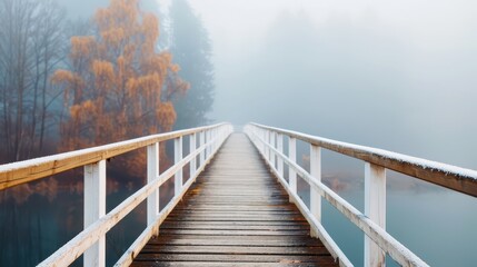 Naklejka premium A mist-covered bridge leads through a foggy landscape flanked by autumn trees, creating a picturesque scene filled with seasonal charm and a sense of mystery.