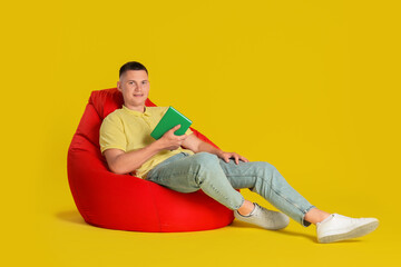 Handsome man with book on red bean bag chair against yellow background