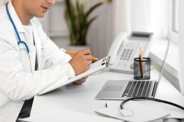 Male doctor with clipboard working at table in clinic, closeup