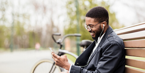 An African American businessman is using his phone while sitting on a park bench with his bicycle nearby. The mood is relaxed and professional, set in a green, serene park.