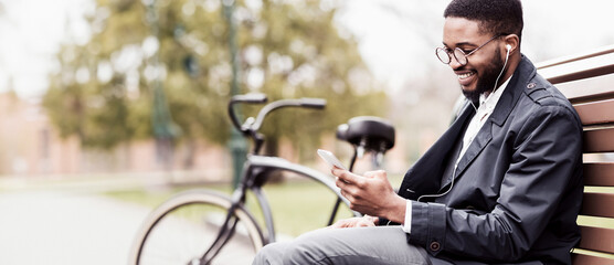 A professional black man is sitting in a park with his phone and bicycle beside him. The mood is contemporary and relaxed, reflecting a modern lifestyle in a natural setting.