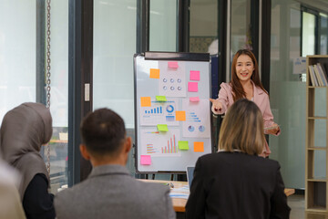 A cheerful and confident Asian businesswoman stands and presents bar chart data from a projector screen to her office colleague's Asian business leader role at the meeting.