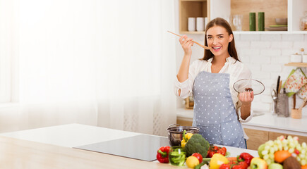 A young woman smiles in her kitchen, wearing an apron and holding a wooden spoon, while fresh produce sits on the counter around her.